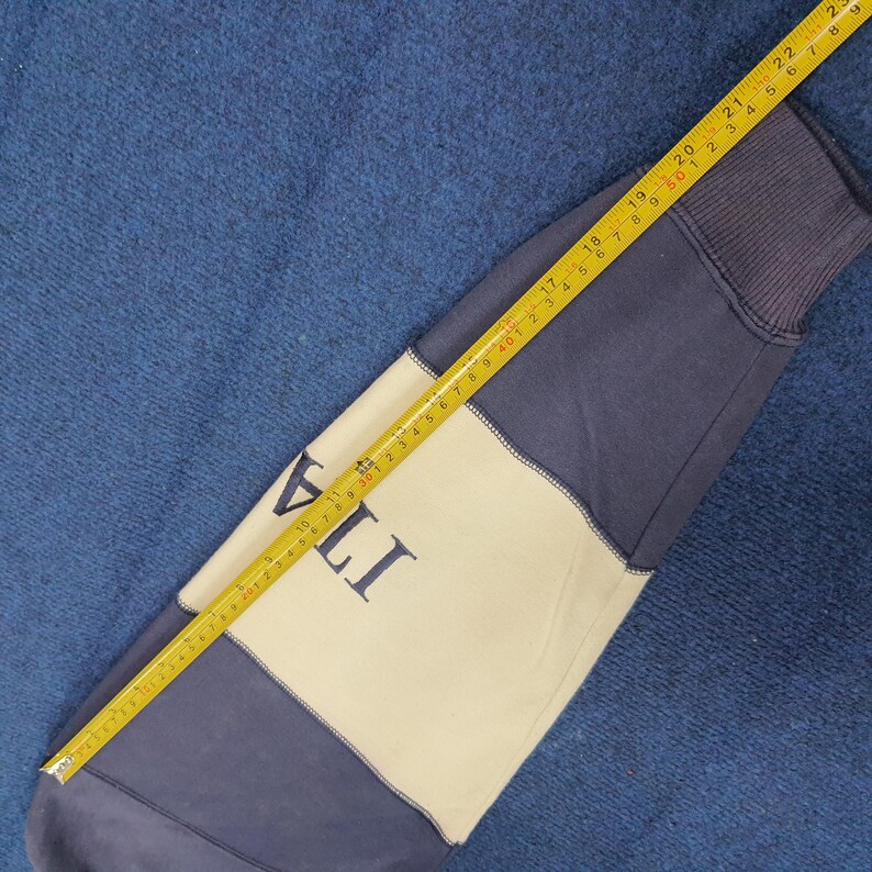 Puede incluir: Una manga de sudadera azul marino y crema con la palabra "ITA" impresa en la secci&oacute;n crema. Una cinta m&eacute;trica amarilla se coloca sobre la manga, mostrando medidas en cent&iacute;metros.