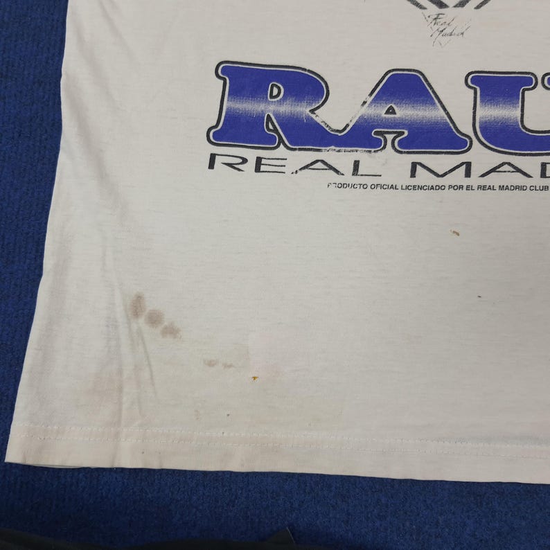 May include: White t-shirt with the word "RAUL" in blue block letters. The text "REAL MADRID" is below the name. The shirt has a stain on the lower left side and the text "PRODUCTO OFICIAL LICENCIADO POR EL REAL MADRID CLUB" is printed below.