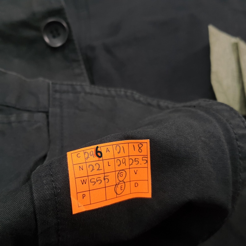 May include: Close-up of a dark green garment with a button and a small orange tag. The tag has black text and numbers, including "C 296", "A 21 18", "N 22", "L 29 25.5", and "W S55".