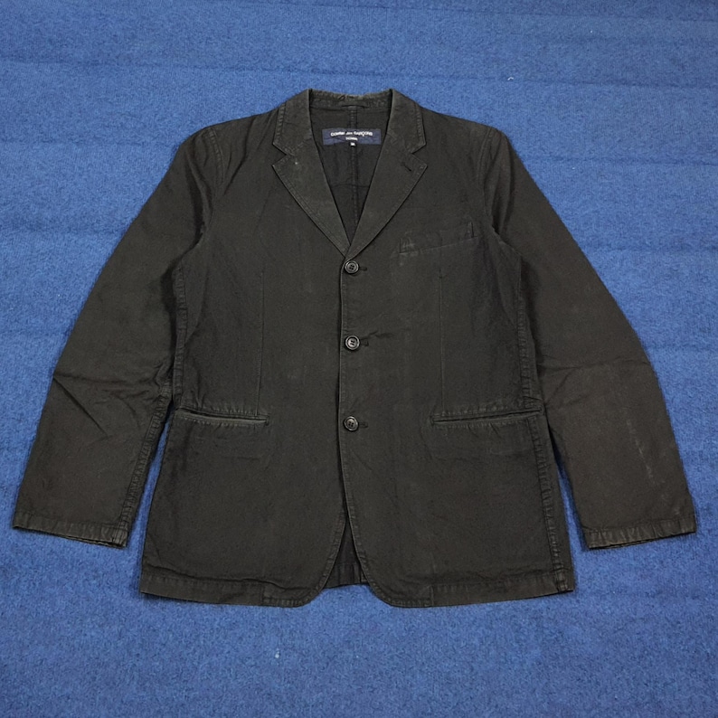 May include: A black blazer with three buttons and two front pockets. The blazer has a notched lapel and long sleeves. The label on the inside of the blazer is visible.