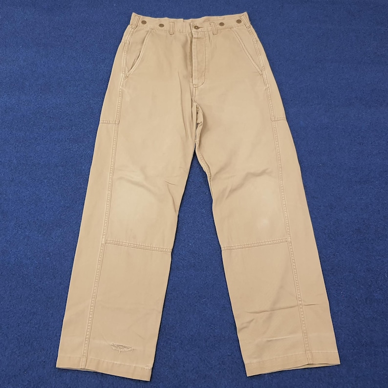 May include: Khaki-colored work pants with a straight leg design. The pants feature multiple pockets, reinforced knee patches, and button closures at the waist. The pants are made of a durable fabric and are suitable for work or casual wear.