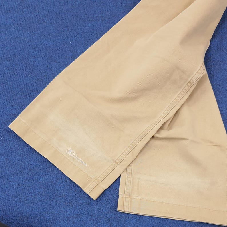 May include: Tan-colored pants with a wide leg design. The pants have visible stitching along the seams and a small embroidered detail near the hem. The fabric appears to be a sturdy cotton or blend, suitable for casual wear.