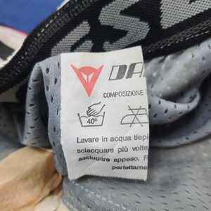 Puede incluir: Primer plano de una etiqueta blanca con la marca "DA" y un logotipo rojo. La etiqueta incluye instrucciones de lavado, como un s&iacute;mbolo de lavado a mano a 40 grados. La etiqueta est&aacute; adherida a una tela gris perforada.