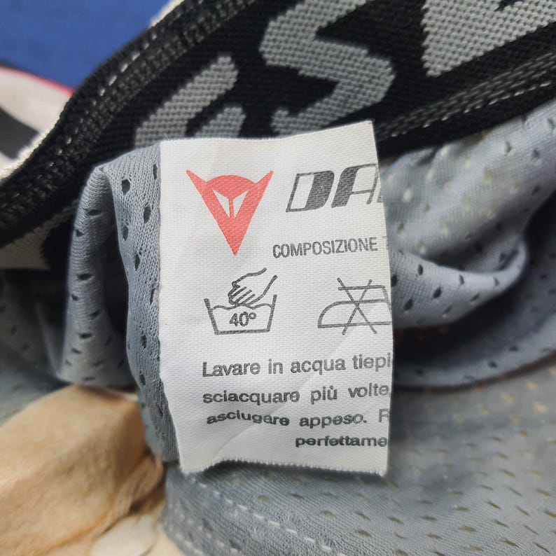 Puede incluir: Primer plano de una etiqueta blanca con la marca "DA" y un logotipo rojo. La etiqueta incluye instrucciones de lavado, como un s&iacute;mbolo de lavado a mano a 40 grados. La etiqueta est&aacute; adherida a una tela gris perforada.