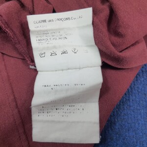 May include: Close-up of a burgundy fabric with a white tag attached. The tag features the text "COMME des GAR&Ccedil;ONS Co. Ltd." and care instructions. The fabric appears to be cotton.