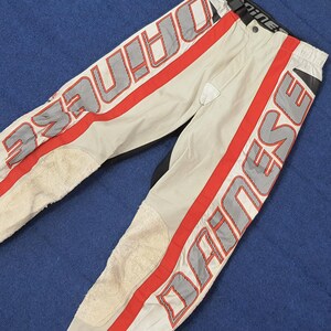 Puede incluir: Pantalones de moto color crema con rayas rojas y la palabra "DAINESE" en letras grandes y contorneadas. Los pantalones tienen refuerzos en las rodillas y detalles en negro. Dise&ntilde;ados para motociclismo.