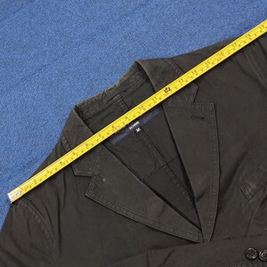 May include: Dark olive green blazer with a notched lapel and two-button closure. A yellow measuring tape is laid across the jacket. The jacket has the word "HOMME" on the label. The blazer is size M.