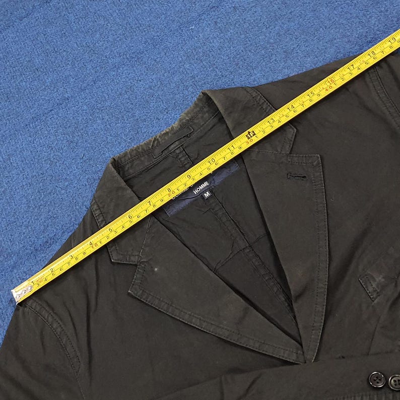 May include: Dark olive green blazer with a notched lapel and two-button closure. A yellow measuring tape is laid across the jacket. The jacket has the word "HOMME" on the label. The blazer is size M.