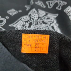 K&ouml;nnte beinhalten: Nahaufnahme eines schwarzen Kleidungsst&uuml;cks mit einem wei&szlig;en Grafikdesign und dem Text "U.S. NAVY". Ein orangefarbenes Etikett mit schwarzem Text und Zahlen ist am Kleidungsst&uuml;ck befestigt.