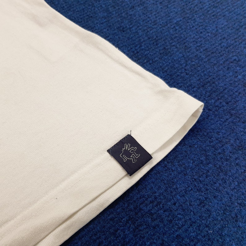 Puede incluir: Camiseta blanca con una etiqueta negra que presenta un gr&aacute;fico estilizado de figuras bailando.