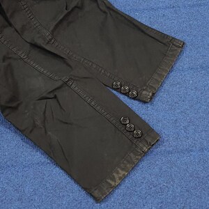 May include: Close-up of black pants legs with three buttons at the cuff. The pants appear to be made of a durable fabric, with visible stitching and a slightly worn appearance. The buttons are dark and round.