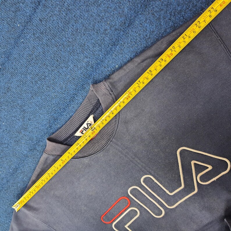 Puede incluir: Sudadera FILA azul marino con logo blanco y rojo. Una cinta m&eacute;trica amarilla est&aacute; colocada sobre la sudadera. La sudadera tiene cuello redondo y mangas largas. El logo est&aacute; delineado en blanco con un acento rojo.