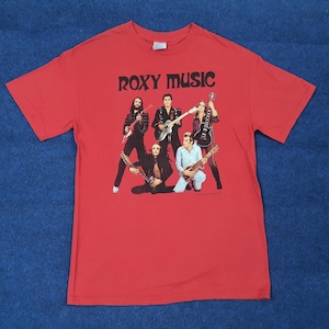 May include: Red t-shirt featuring the band Roxy Music with a graphic of the band members playing guitars. The band's name is printed in black block letters across the top. The shirt is made of a soft material.