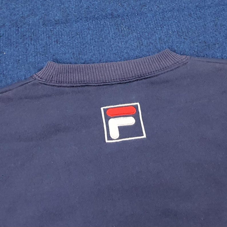 Puede incluir: Sudadera azul marino con cuello de canal&eacute;. La sudadera presenta un logotipo cuadrado blanco con un dise&ntilde;o "F" y "L" rojo y blanco. El fondo es una superficie azul texturizada.