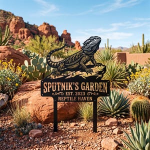 Puede incluir: Un letrero de jardín de metal negro con la silueta de un dragón barbudo en la parte superior. El letrero muestra el texto "SPUTNIK'S GARDEN", "EST. 2023" y "REPTILE HAVEN" en una fuente clara. Se sostiene mediante dos estacas metálicas.