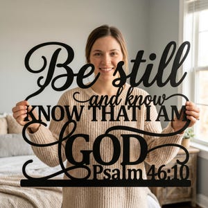 May include: Black metal wall art with the words "Be still and know that I am GOD Psalm 46:10" in a decorative font. The sign is held up by a person in a beige sweater.