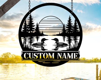 May include: A black metal sign with a circular design, featuring a lake scene with two loons, trees, and a setting sun. The sign hangs from chains and includes the text "CUSTOM NAME" in a rectangular banner.