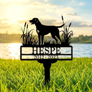 Puede incluir: Estaca de jardín de metal negro con la silueta de un perro, juncos, el nombre "HESPE" y los años "2012-2023". El fondo muestra un lago y una puesta de sol. La estaca está en la hierba.