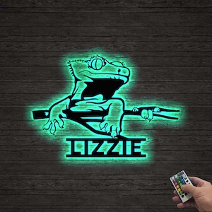 May include: A black metal gecko silhouette with the name "LIZZIE" below, illuminated with a green glow. The gecko is perched on a branch, and a remote control is in the lower right corner. The background is a dark wood-grain pattern.