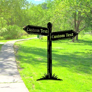 Op de afbeelding: Zwarte metalen wegwijzer met twee pijlen, elk met het opschrift "Custom Text". De wegwijzer staat tegen een achtergrond van groen gras, bomen en een wandelpad.