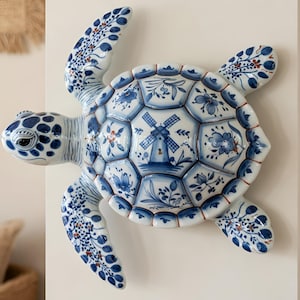 May include: A ceramic turtle wall decor piece. The turtle is white with blue floral and windmill designs. The shell is segmented, and the flippers and head are detailed with similar patterns. The decor is mounted on a white wall.