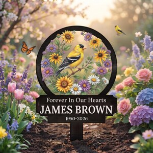 May include: A black circular memorial garden stake featuring a yellow goldfinch perched on a branch surrounded by sunflowers and purple daisies. Below the circle, a rectangular plaque displays the text Forever In Our Hearts, JAMES BROWN, 1950-2026.
