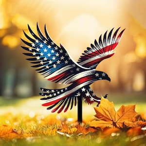Puede incluir: Una estaca de jardín de metal con un águila con las alas extendidas, pintada con el diseño de la bandera estadounidense. El águila es roja, blanca y azul, con estrellas y rayas. La estaca está en un área cubierta de hierba con hojas caídas.