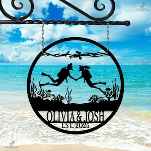 May include: Black metal sign with a circular design depicting two scuba divers holding hands underwater. The sign includes the names "OLIVIA & JOSH" and "EST. 2025", set against a beach and ocean backdrop.