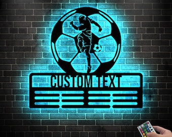 May include: A black metal silhouette of a soccer player kicking a ball inside a soccer ball design. The silhouette is illuminated with blue light. The design has a black metal plaque below with the text "CUSTOM TEXT" and five lines for personalization.