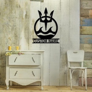 May include: Black metal wall decor featuring a trident with waves and the text "CUSTOM NAME". The decor is mounted on a light wood wall with a white dresser and a white chair in the foreground.