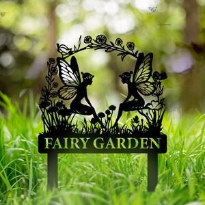 Puede incluir: Estaca de jard&iacute;n de metal negro con dos hadas sentadas en una rama con flores y hojas. La estaca tiene un letrero que dice "FAIRY GARDEN".