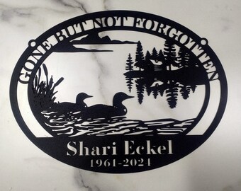 May include: Black metal oval memorial plaque with a silhouette of two loons swimming on a lake with trees reflected in the water. The text "Gone But Not Forgotten" is written around the top edge of the plaque. The text "Shari Eckel 1961-2021" is written below the image.