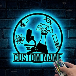 以下が含まれることがあります： 顕微鏡、ビーカー、原子、DNA鎖、電球が付いた脳が描かれた青いネオンサイン。サインには「CUSTOM NAME」と表示され、リモコンが付属しています。
