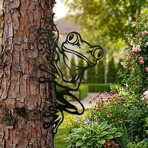 May include: A black metal frog sculpture climbing a tree trunk. The frog has large eyes and is attached to the tree with its hands and feet. The background includes a garden with flowers and greenery.