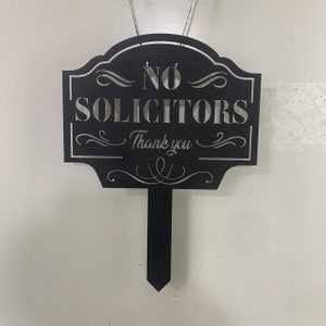 K&ouml;nnte beinhalten: Schwarzes Metallschild mit den Worten "NO SOLICITORS" und "Thank you" in dekorativer Schrift. Das Schild hat einen spitzen Pfahl zur einfachen Platzierung in einem Garten oder Hof.