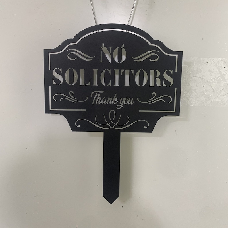 K&ouml;nnte beinhalten: Schwarzes Metallschild mit den Worten "NO SOLICITORS" und "Thank you" in dekorativer Schrift. Das Schild hat einen spitzen Pfahl zur einfachen Platzierung in einem Garten oder Hof.