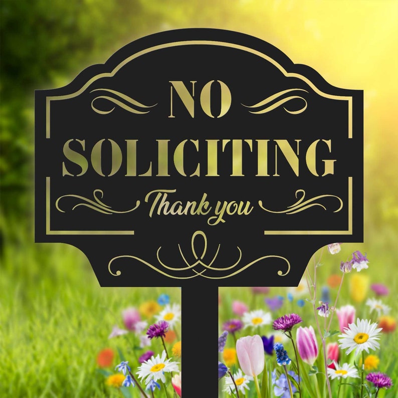 K&ouml;nnte beinhalten: Ein schwarzes Metallschild mit goldfarbenem Schriftzug "NO SOLICITING Thank you". Das Schild hat ein verziertes Design und befindet sich in einem Garten mit bunten Blumen. Das Schild ist an einem schwarzen Pfahl befestigt.