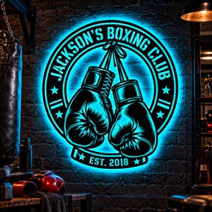 May include: A circular wall sign with bright cyan LED backlighting featuring a graphic of two tied boxing gloves in the center. The text reads JACKSON'S BOXING CLUB and EST. 2018. The design is black with a bold, glowing cyan outline.