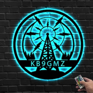 Pode incluir: Arte de parede em metal preto com iluminação LED azul. Apresenta um design de torre de rádio com as letras KB9GMZ. A obra é circular, com uma borda preta, e está montada numa parede de tijolo escuro.