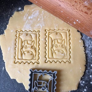 May include: A light-coloured dough with two Totoro-shaped cookie cutouts and a grey stamp-shaped cookie cutter. A wooden rolling pin rests on the dough. The cookie cutouts are in the shape of a postage stamp.