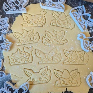 Peut inclure: Gros plan sur une feuille de pâte à biscuits avec divers emporte-pièces en forme de personnages Pokémon. Les emporte-pièces sont en plastique blanc et la pâte est de couleur brun clair.