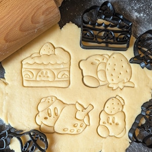 Puede incluir: Un rodillo de madera descansa sobre una masa con varias formas de cortadores de galletas presionadas. Las formas incluyen una rebanada de pastel, una fresa y otros personajes de dibujos animados. También se ven cortadores de galletas negros.