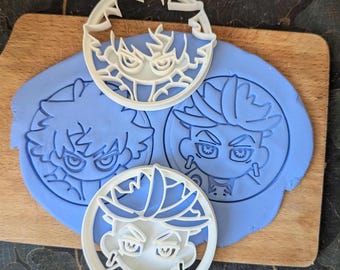 Op de afbeelding: Witte plastic koekjessnijders in de vorm van anime-achtige gezichten worden getoond op een houten snijplank. De snijders zijn geplaatst op blauw deeg, met afdrukken van de gezichten zichtbaar. De snijders hebben gedetailleerde gelaatstrekken.
