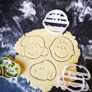 May include: White plastic cookie cutters in the shapes of Charlie Brown, Snoopy, Lucy, and Linus from the Peanuts cartoon. The cutters are on a black surface with a sheet of cookie dough underneath.