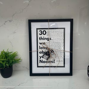 May include: A black framed print with the text "30 things we love about you Naomi!". The print is tied with twine and a silver charm. A small potted plant is on the left. The print is a personalized gift.