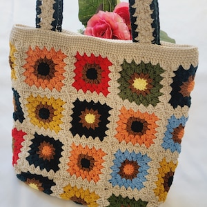 May include: A crocheted tote bag with a colorful granny square pattern. The bag has two long straps and is made with a beige yarn. The squares are in shades of red, orange, yellow, green, blue, and brown.