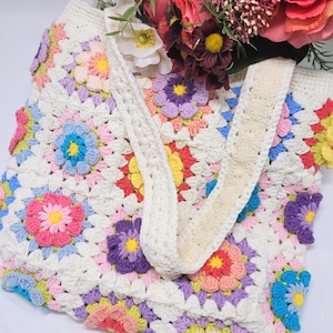May include: A white crocheted tote bag with a colorful floral pattern. The bag has a wide, white strap and is made of granny squares.