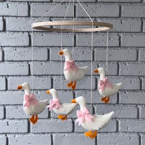 May include: A baby mobile featuring five white felt geese with orange beaks and feet, each adorned with a pink bow. The geese are suspended from a wooden ring and a geometric wooden piece, hanging against a gray brick wall.