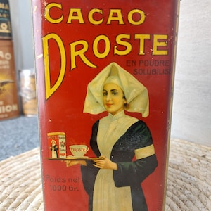 Puede incluir: Una lata de metal roja rectangular con el texto CACAO DROSTE EN POUDRE SOLUBILISE. El frente muestra la ilustración de una persona con tocado blanco y ropa oscura sosteniendo una bandeja con una taza y una lata pequeña. Poids net 1000 Gr.