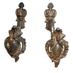Peut inclure: Une paire d'appliques murales assorties en métal doré vieilli. Chaque applique présente un design orné et symétrique avec des volutes complexes, des motifs en forme de feuilles et un élément central en forme de goutte, avec une finition patinée.